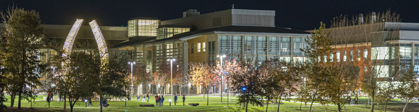 UC Merced at night with illuminated buildings, students walking, and a lit Beginnings Scupture on the Quad